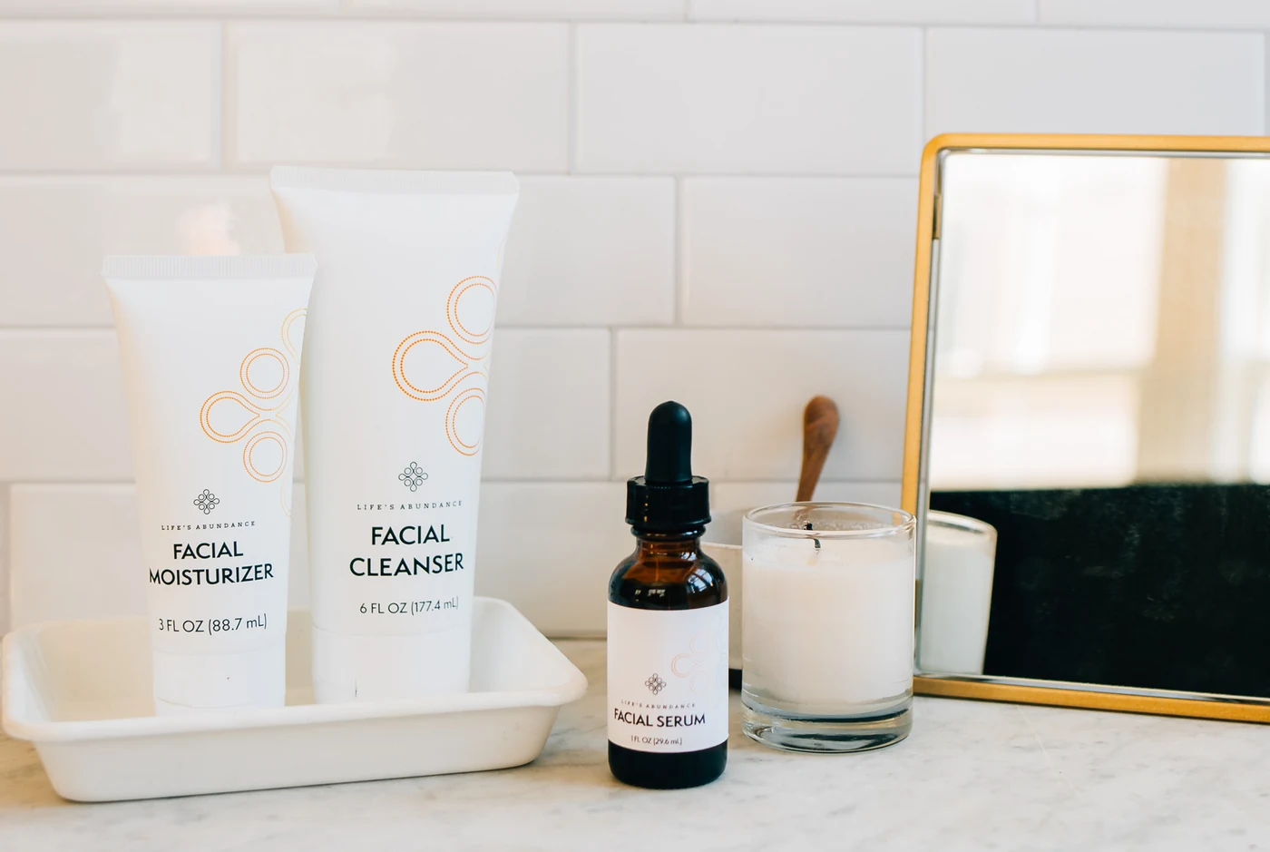 Life’s Abundance facial cleanser, moisturizer and serum beside a candle and mirror on a marble counter